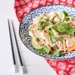 A colourful arrangement of vibrant herbs, snow peas, thinly sliced cucumbers, shredded chicken, julienned carrots, and chili mixed with rice noodles is displayed on a blue and white plate atop a red oriental tablecloth, accompanied by chopsticks.