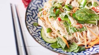 A colourful arrangement of vibrant herbs, snow peas, thinly sliced cucumbers, shredded chicken, julienned carrots, and chili mixed with rice noodles is displayed on a blue and white plate atop a red oriental tablecloth, accompanied by chopsticks.