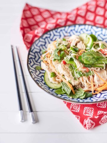 A colourful arrangement of vibrant herbs, snow peas, thinly sliced cucumbers, shredded chicken, julienned carrots, and chili mixed with rice noodles is displayed on a blue and white plate atop a red oriental tablecloth, accompanied by chopsticks.