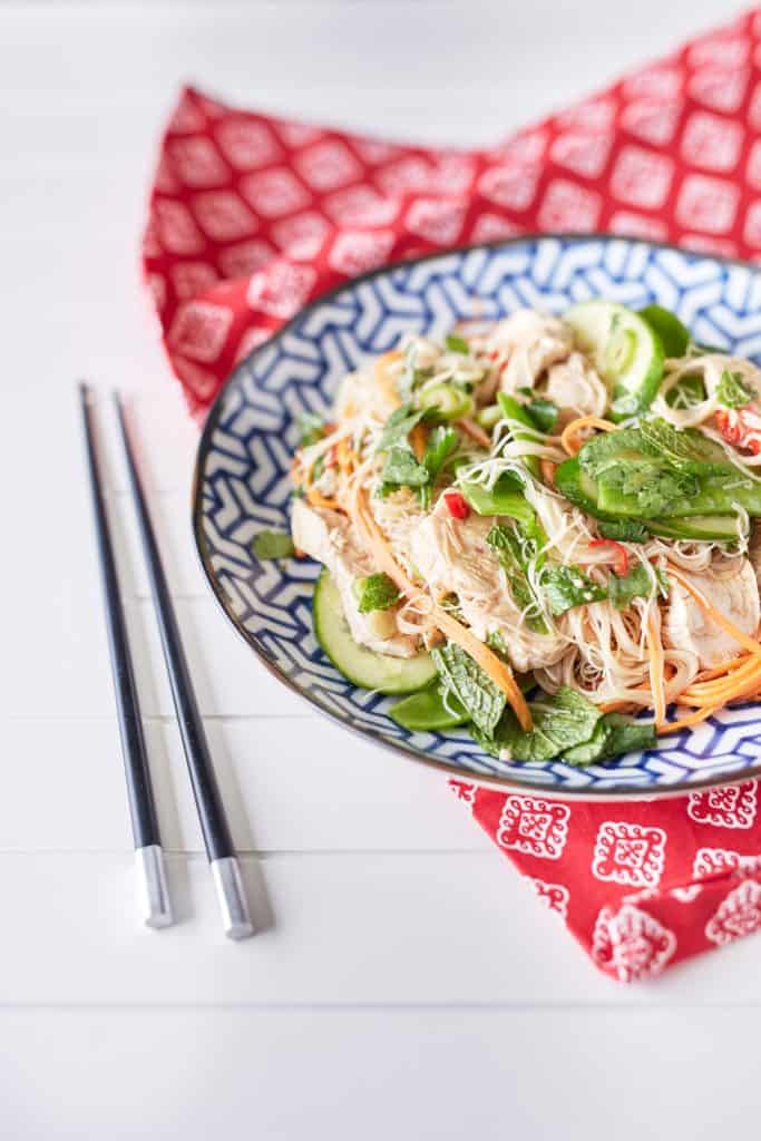 A colourful arrangement of vibrant herbs, snow peas, thinly sliced cucumbers, shredded chicken, julienned carrots, and chili mixed with rice noodles is displayed on a blue and white plate atop a red oriental tablecloth, accompanied by chopsticks.