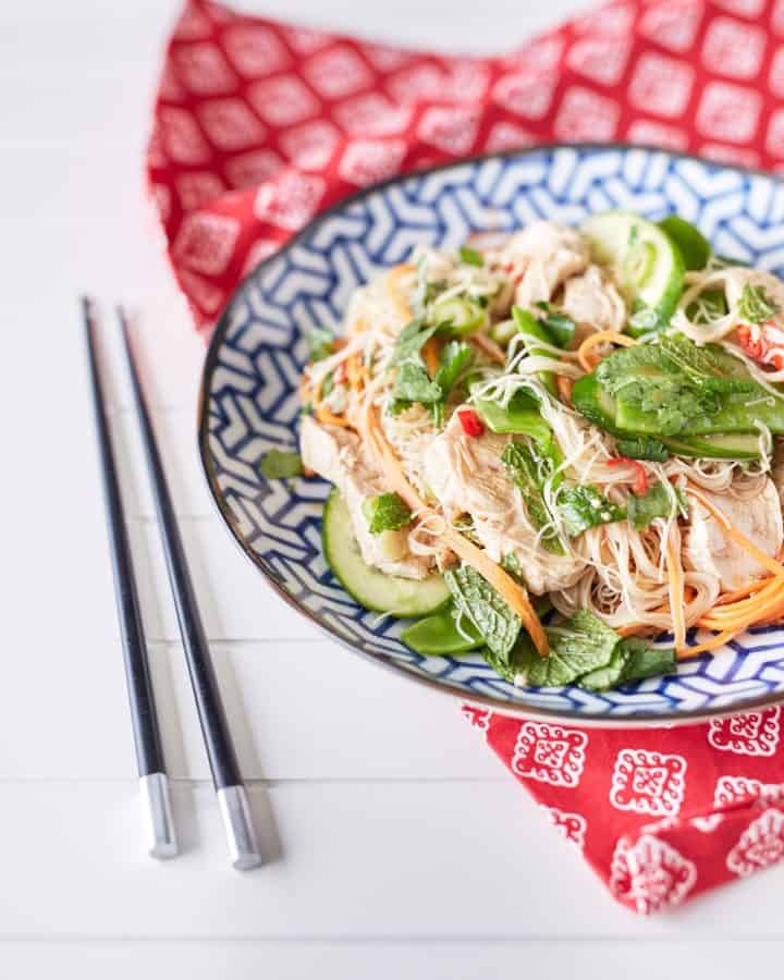 A colourful arrangement of vibrant herbs, snow peas, thinly sliced cucumbers, shredded chicken, julienned carrots, and chili mixed with rice noodles is displayed on a blue and white plate atop a red oriental tablecloth, accompanied by chopsticks.