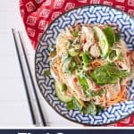 A colourful arrangement of vibrant herbs, snow peas, thinly sliced cucumbers, shredded chicken, julienned carrots, and chili mixed with rice noodles is displayed on a blue and white plate atop a red oriental tablecloth, accompanied by chopsticks.