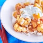 A vibrant cauliflower and pumpkin curry in a deep yellow-orange sauce, topped with white yogurt speckled with cucumber, served on a bed of rice, plated on a white dish, placed on a blue tablecloth.