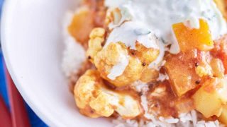 A vibrant cauliflower and pumpkin curry in a deep yellow-orange sauce, topped with white yogurt speckled with cucumber, served on a bed of rice, plated on a white dish, placed on a blue tablecloth.