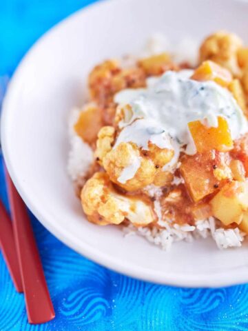 A vibrant cauliflower and pumpkin curry in a deep yellow-orange sauce, topped with white yogurt speckled with cucumber, served on a bed of rice, plated on a white dish, placed on a blue tablecloth.