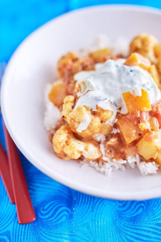 A vibrant cauliflower and pumpkin curry in a deep yellow-orange sauce, topped with white yogurt speckled with cucumber, served on a bed of rice, plated on a white dish, placed on a blue tablecloth.