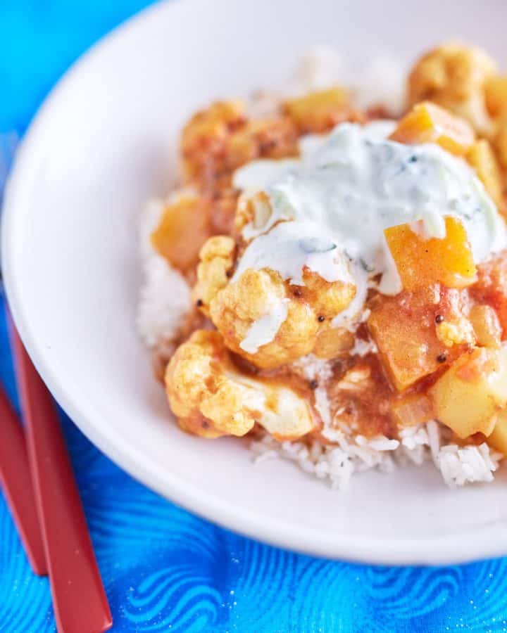 A vibrant cauliflower and pumpkin curry in a deep yellow-orange sauce, topped with white yogurt speckled with cucumber, served on a bed of rice, plated on a white dish, placed on a blue tablecloth.