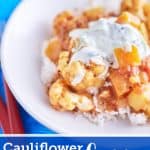 A vibrant cauliflower and pumpkin curry in a deep yellow-orange sauce, topped with white yogurt speckled with cucumber, served on a bed of rice, plated on a white dish, placed on a blue tablecloth.