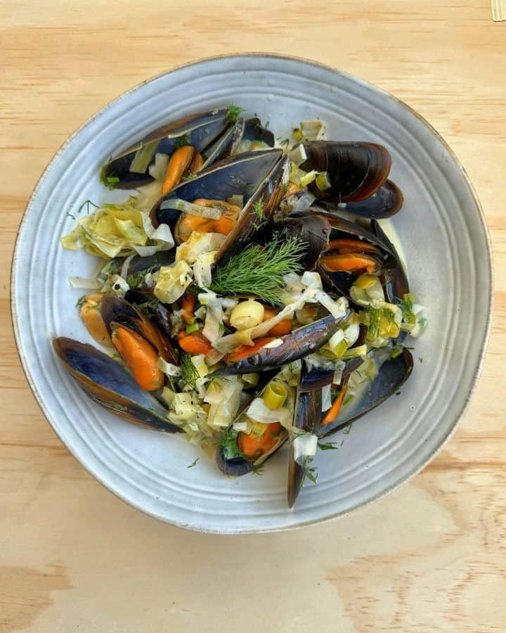 Plump mussels in cream sauce, still in their shells, served on a pale blue plate with leeks and garnished with fresh dill.