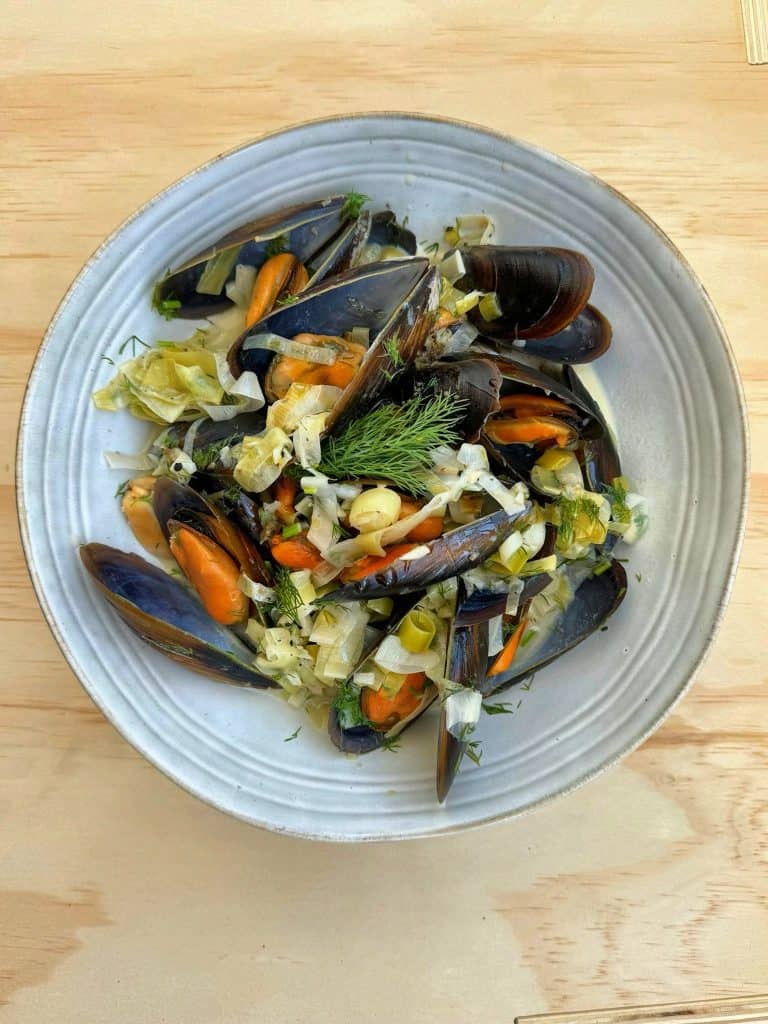 Plump mussels in cream sauce, still in their shells, served on a pale blue plate with leeks and garnished with fennel leaves.