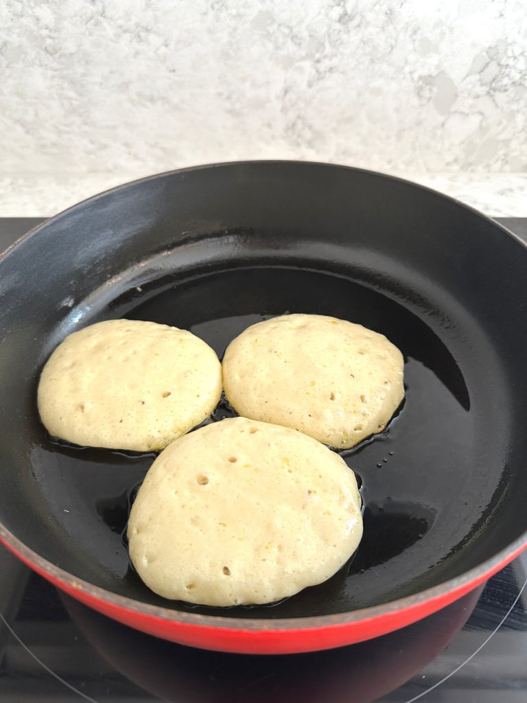 Three protein buttermilk pancakes cooking in a skillet with bubbles appearing on the surface.