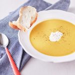 Bright yellow pumpkin and carrot soup garnished with pepper in a white bowl, served with a side of crusty bread on a blue tablecloth.