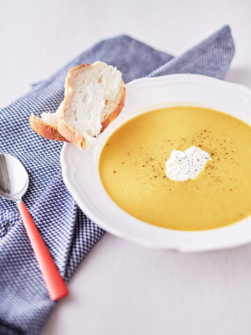 Bright yellow pumpkin and carrot soup garnished with pepper in a white bowl, served with a side of crusty bread on a blue tablecloth.