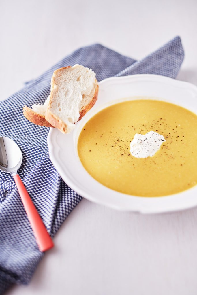 Bright yellow pumpkin and carrot soup garnished with pepper in a white bowl, served with a side of crusty bread on a blue tablecloth.