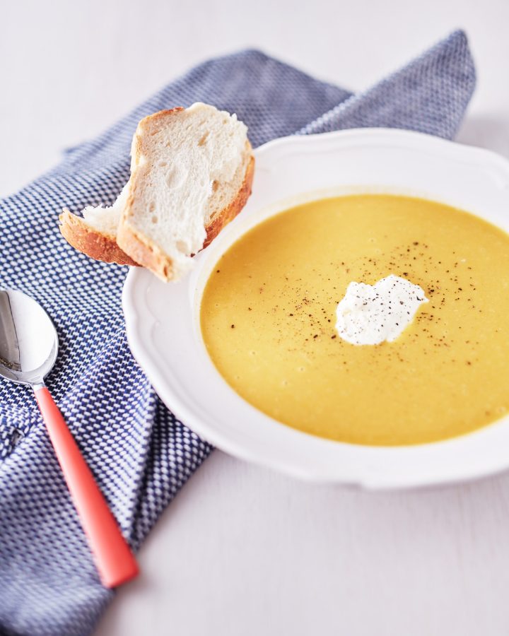 Bright yellow pumpkin and carrot soup garnished with pepper in a white bowl, served with a side of crusty bread on a blue tablecloth.