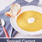 Bright yellow pumpkin and carrot soup garnished with pepper in a white bowl, served with a side of crusty bread on a blue tablecloth.