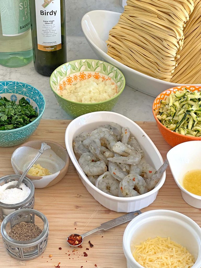 All the ingredients for this courgette prawn pasta are laid out pre-measured and ready to use, including prawns, onion, pasta, wine, and grated courgette.