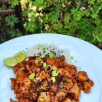 Sticky honey garlic ginger chicken pieces, garnished with scallions, black sesame seeds, and a wedge of lime, are arranged on a white plate.