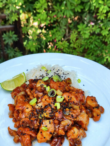 Sticky honey garlic ginger chicken pieces, garnished with scallions, black sesame seeds, and a wedge of lime, are arranged on a white plate.