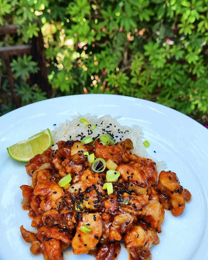 Sticky honey garlic ginger chicken pieces, garnished with scallions, black sesame seeds, and a wedge of lime, are arranged on a white plate.