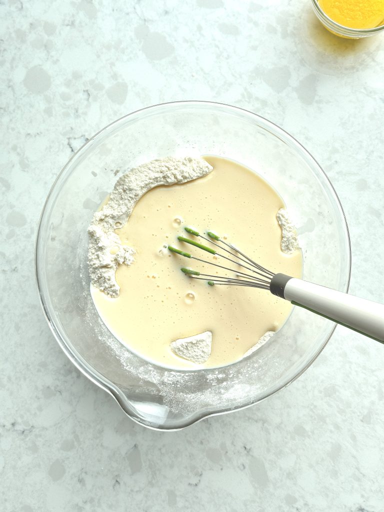 A glass bowl containing wet ingredients added to dry ingredients but not mixed, with a whisk resting on the bowl and a small bowl with melted butter beside it.