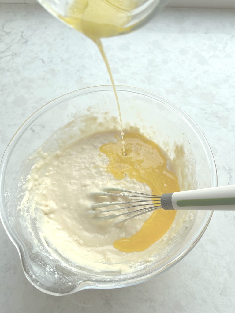 Melted butter being added to batter in a glass bowl while whisking.