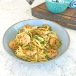 Courgette prawn pasta, served in a blue bowl on a marble table, features linguini topped with caramelised prawns, courgette, and parsley, accompanied by a wine glass.