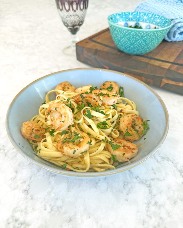 Courgette prawn pasta, served in a blue bowl on a marble table, features linguini topped with caramelised prawns, courgette, and parsley, accompanied by a wine glass.