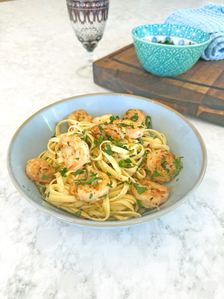 Courgette prawn pasta, served in a blue bowl on a marble table, features linguini topped with caramelised prawns, courgette, and parsley, accompanied by a wine glass.