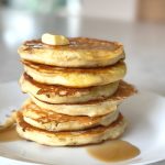 Six fluffy buttermilk pancakes stacked on top of each other with melting butter and drizzled maple syrup on a white plate.