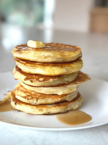 Six fluffy buttermilk pancakes stacked on top of each other with melting butter and drizzled maple syrup on a white plate.