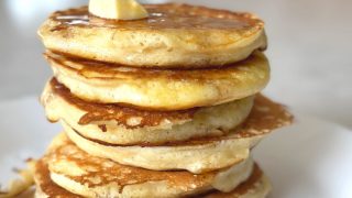 Six fluffy buttermilk pancakes stacked on top of each other with melting butter and drizzled maple syrup on a white plate.