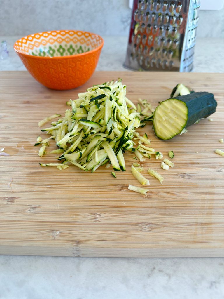 On the chopping board, there is a zucchini partially grated, with one half remaining whole and the other half grated, accompanied by a grater, with an orange bowl visible in the background.