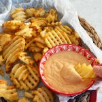 A rustic plate of waffle fries with a bowl of honey BBQ aioli on the side and a hand dipping a fry into the sauce.