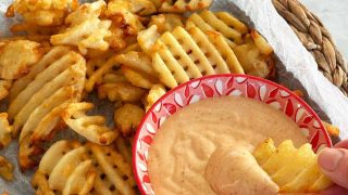 A rustic plate of waffle fries with a bowl of honey BBQ aioli on the side and a hand dipping a fry into the sauce.