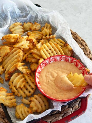 A rustic plate of waffle fries with a bowl of honey BBQ aioli on the side and a hand dipping a fry into the sauce.