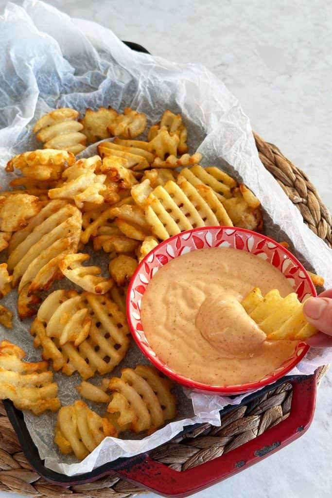 A rustic plate of waffle fries with a bowl of honey BBQ aioli on the side and a hand dipping a fry into the sauce.