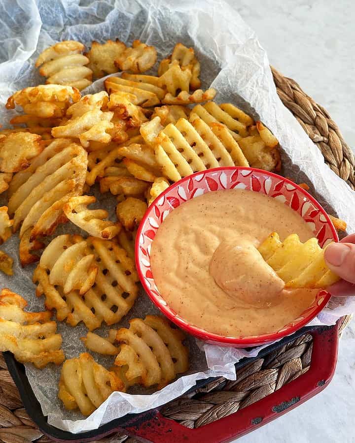 A rustic plate of waffle fries with a bowl of honey BBQ aioli on the side and a hand dipping a fry into the sauce.