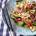 Thai Beef Noodle Salad on a light blue plate with peanuts, cherry tomatoes, cucumbers, snow peas, fresh herbs, and beef on a blue and white tablecloth