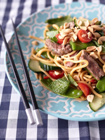 Thai Beef Noodle Salad on a light blue plate with peanuts, cherry tomatoes, cucumbers, snow peas, fresh herbs, and beef on a blue and white tablecloth