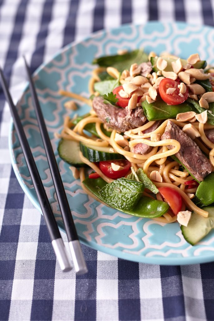 Thai Beef Noodle Salad on a light blue plate with peanuts, cherry tomatoes, cucumbers, snow peas, fresh herbs, and beef on a blue and white tablecloth