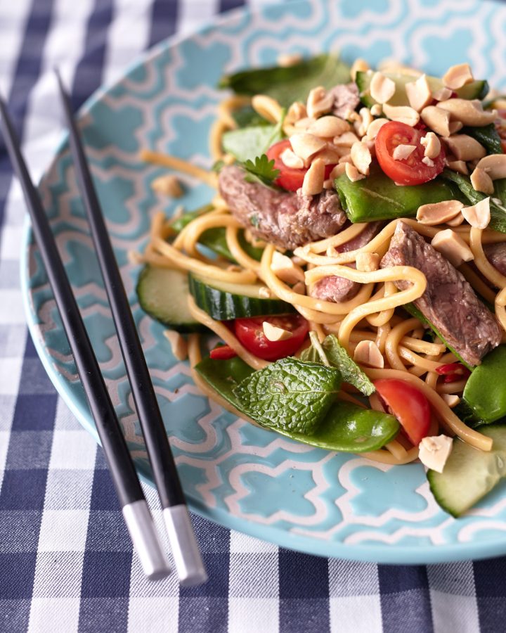 Thai Beef Noodle Salad on a light blue plate with peanuts, cherry tomatoes, cucumbers, snow peas, fresh herbs, and beef on a blue and white tablecloth