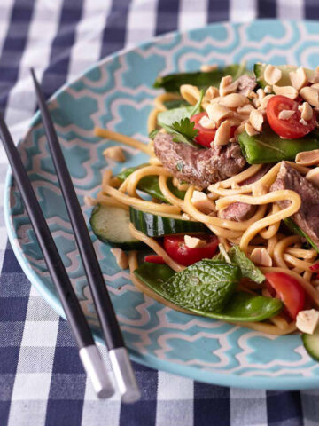 Thai Beef Noodle Salad on a light blue plate with peanuts, cherry tomatoes, cucumbers, snow peas, fresh herbs, and beef on a blue and white tablecloth