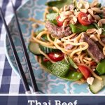 Thai Beef Noodle Salad on a light blue plate with peanuts, cherry tomatoes, cucumbers, snow peas, fresh herbs, and beef on a blue and white tablecloth