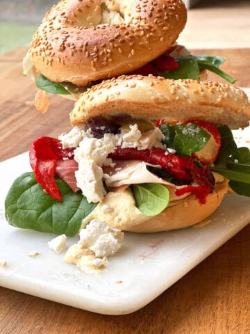 Two bagels with hummus stacked one behind the other, each topped with layers of spinach leaves, feta cheese, roasted red peppers, Serrano ham, and mixed olives.