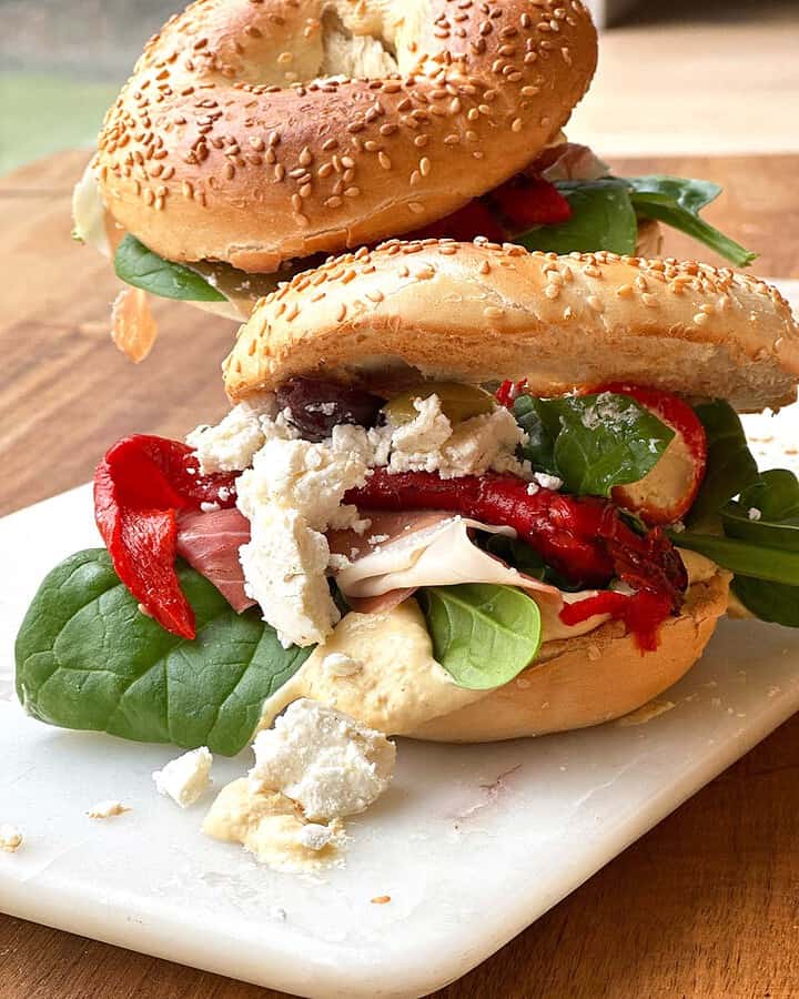 Two bagels with hummus stacked one behind the other, each topped with layers of spinach leaves, feta cheese, roasted red peppers, Serrano ham, and mixed olives.