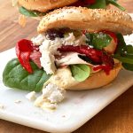 Two bagels with hummus stacked one behind the other, each topped with layers of spinach leaves, feta cheese, roasted red peppers, Serrano ham, and mixed olives.