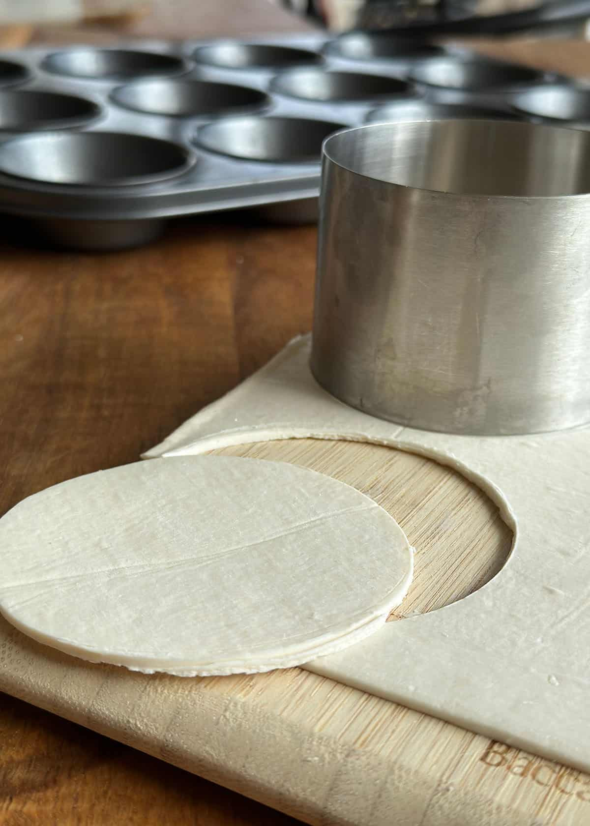 Round piece of puff pastry cut out with a steel cutter on a wooden board, with a muffin tin in the background to make tartlets.