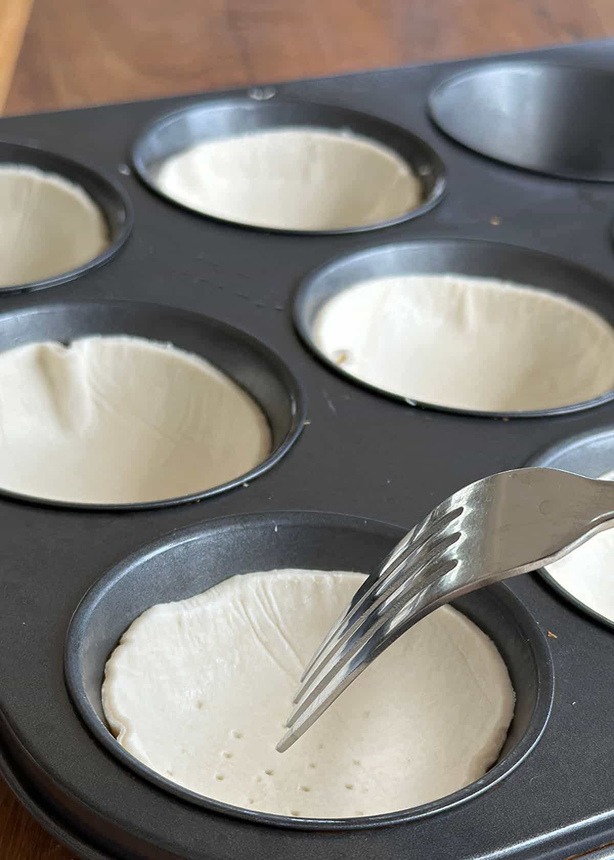 A fork is used to prick the surface of puff pastry pressed into muffin tin cups to prevent puffing during baking.