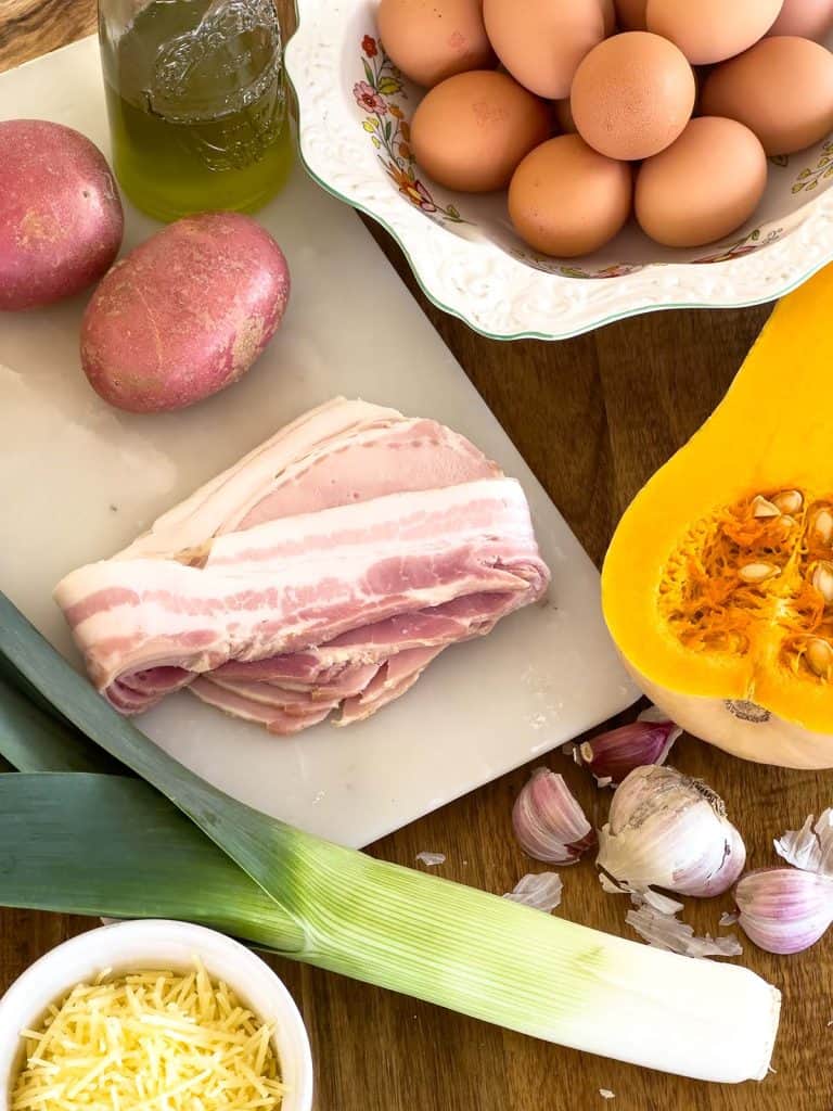 All the ingredients for pumpkin frittata arranged on a wooden board, including pumpkin, leek, bacon, parmesan cheese, oil, eggs, garlic, and potato.